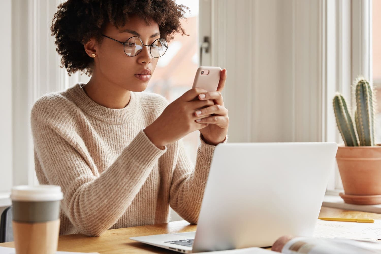 Consórcio: uma mulher de óculos utiliza o celular. Na mesa há um notebook, um copo de café e um cacto.
