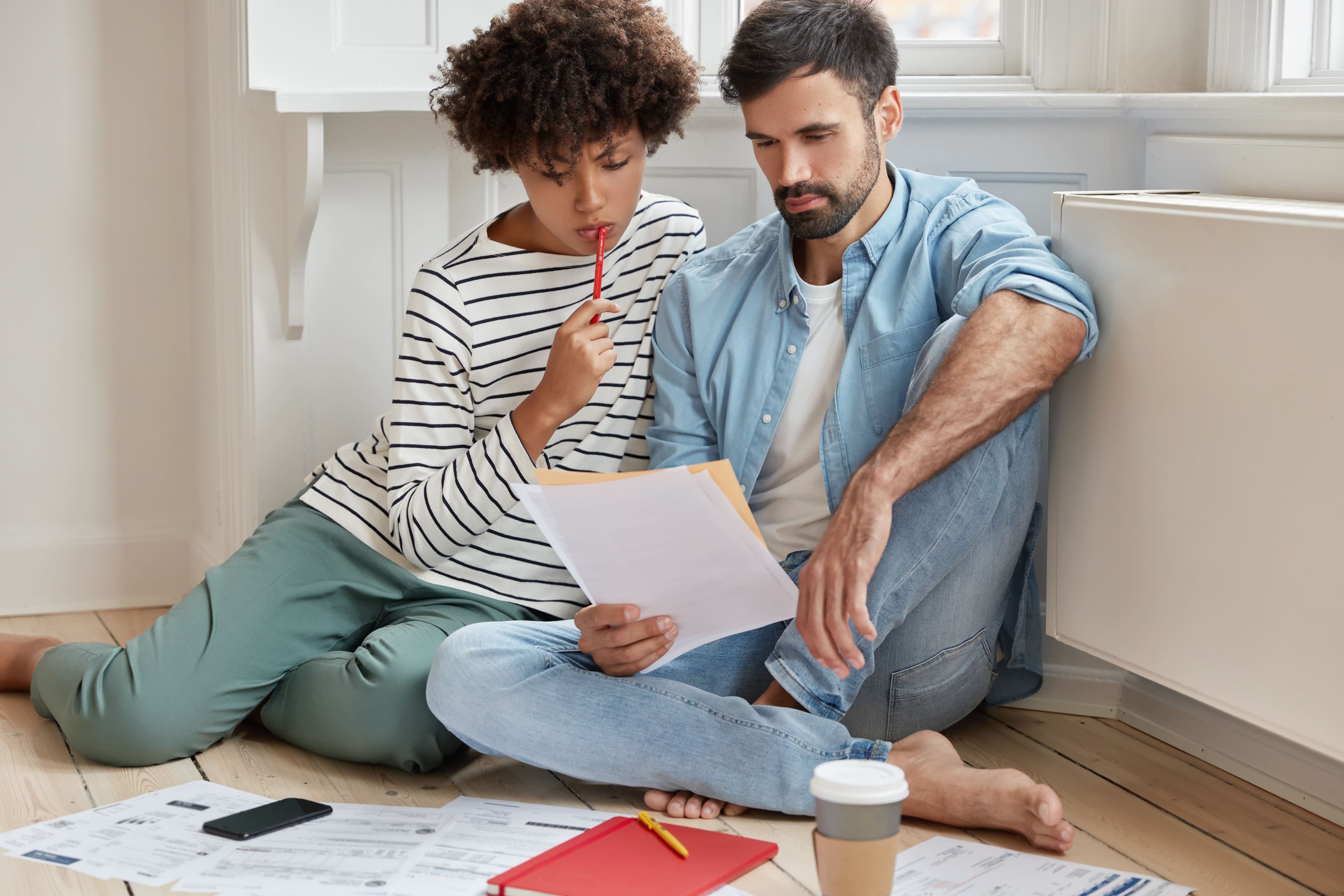Um casal composto por um homem e uma mulher está sentado no chão e lendo papéis, para pensar em uma forma de juntar dinheiro para casar