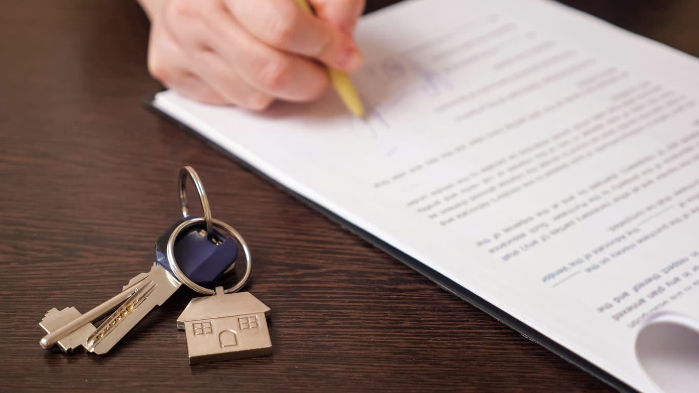 Empréstimo com garantia: uma pessoa está assinando um contrato, ao lado há uma chave com um chaveiro de casa.