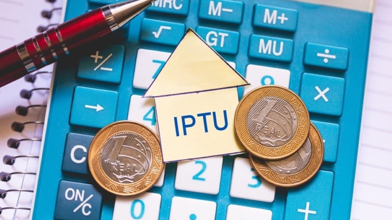 IPTU: imagem mostra uma calculadora em cima de uma caderno. Em cima da calculadora há uma caneta, três moedas de um real e um recorte de papel em formato de casa, com a sigla IPTU escrita dentro.