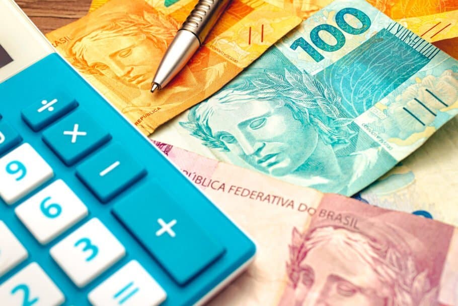 Desenrola Brasil: algumas notas de dinheiro estão espalhadas. Em cima há uma caneta e uma calculadora azul com branco.