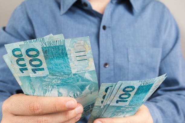Aposentadoria: um homem de camisa social azul está contando notas de cem reais.