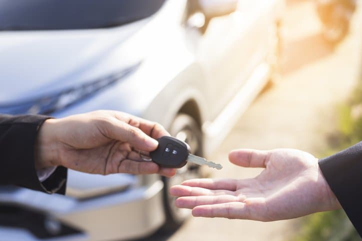 Comprar ou alugar carro: uma pessoa entrega a chave de um veículo a outra pessoa. Ao fundo, há um automóvel.