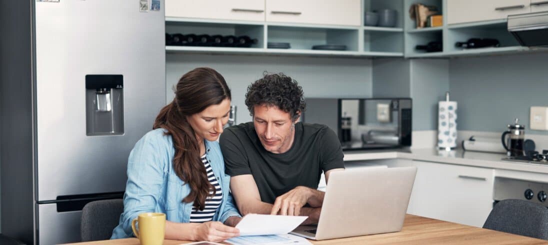 Um casal está sentado à mesa da cozinha, analisando opções de seguro para caminhonete. Na mesa há um notebook e papéis.
