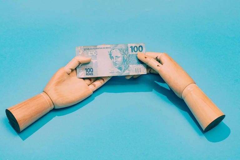 Desenrola Brasil: duas mãos de madeira seguram uma nota de cem reais. O fundo da imagem é azul.