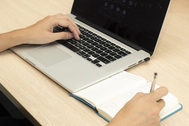 Imposto de Renda: uma pessoa mexe no notebook com a mão esquerda, enquanto faz anotações em um caderno com a mão direita