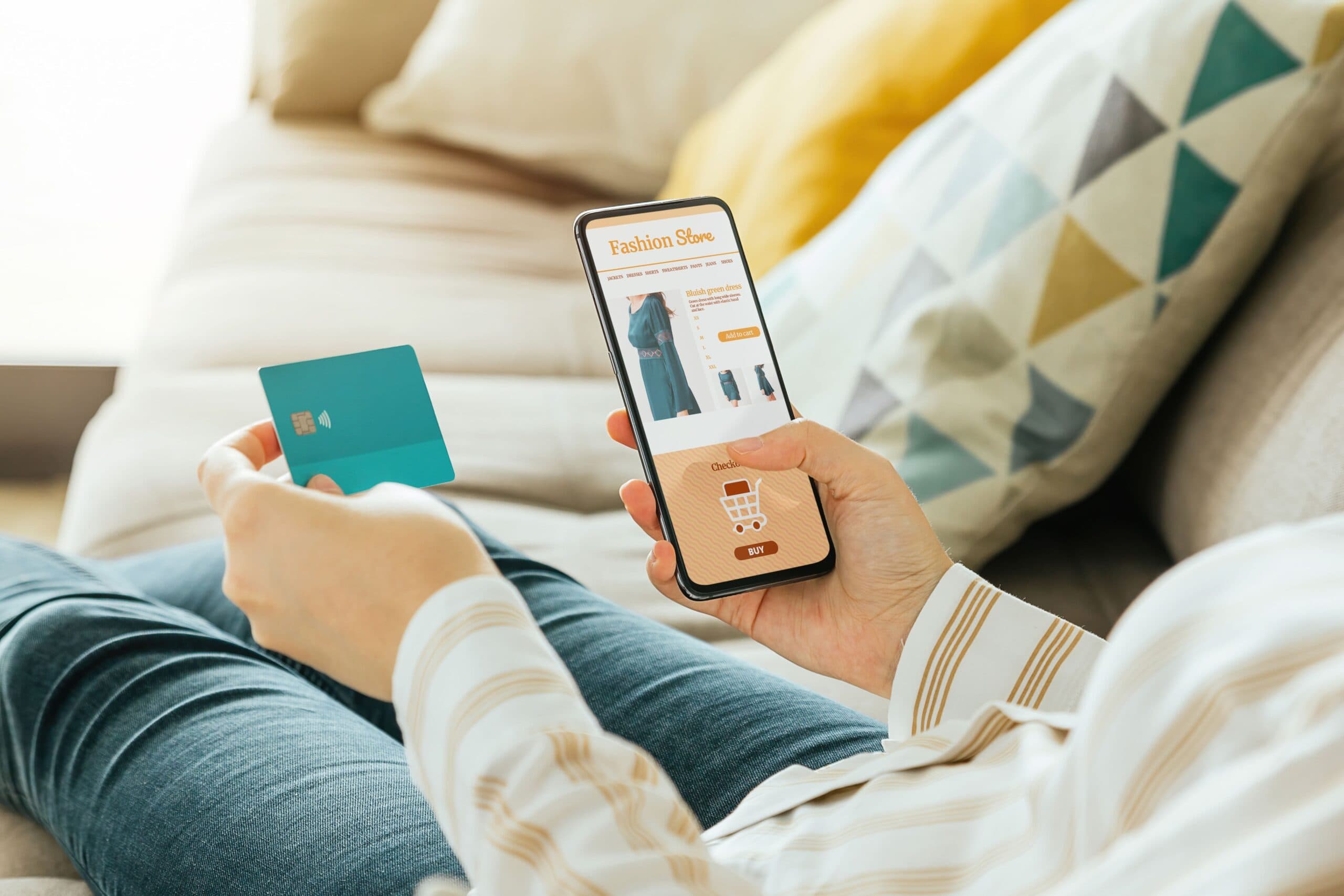 Cashback em cartões de crédito: uma mulher está sentada no sofá, utilizando o cartão para efetuar uma compra online pelo celular.