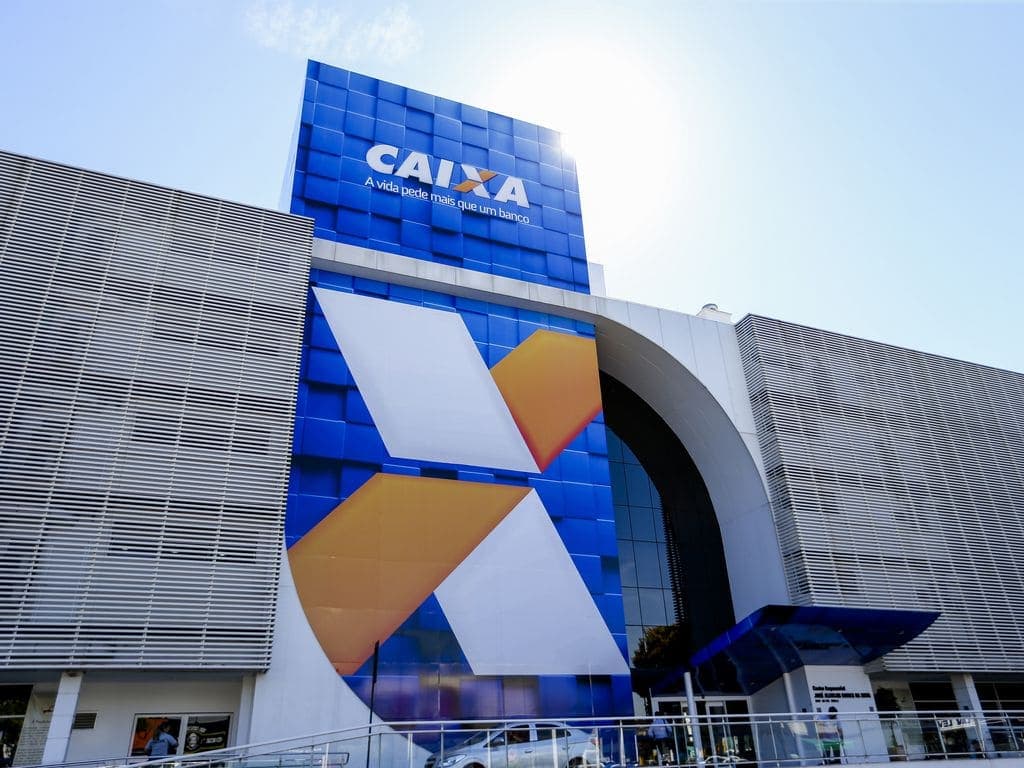 Senha: prédio da Caixa Econômica Federal possui o logo estampado na fachada, acima está escrito Caixa e na entrada há pessoas e um carro estacionado.