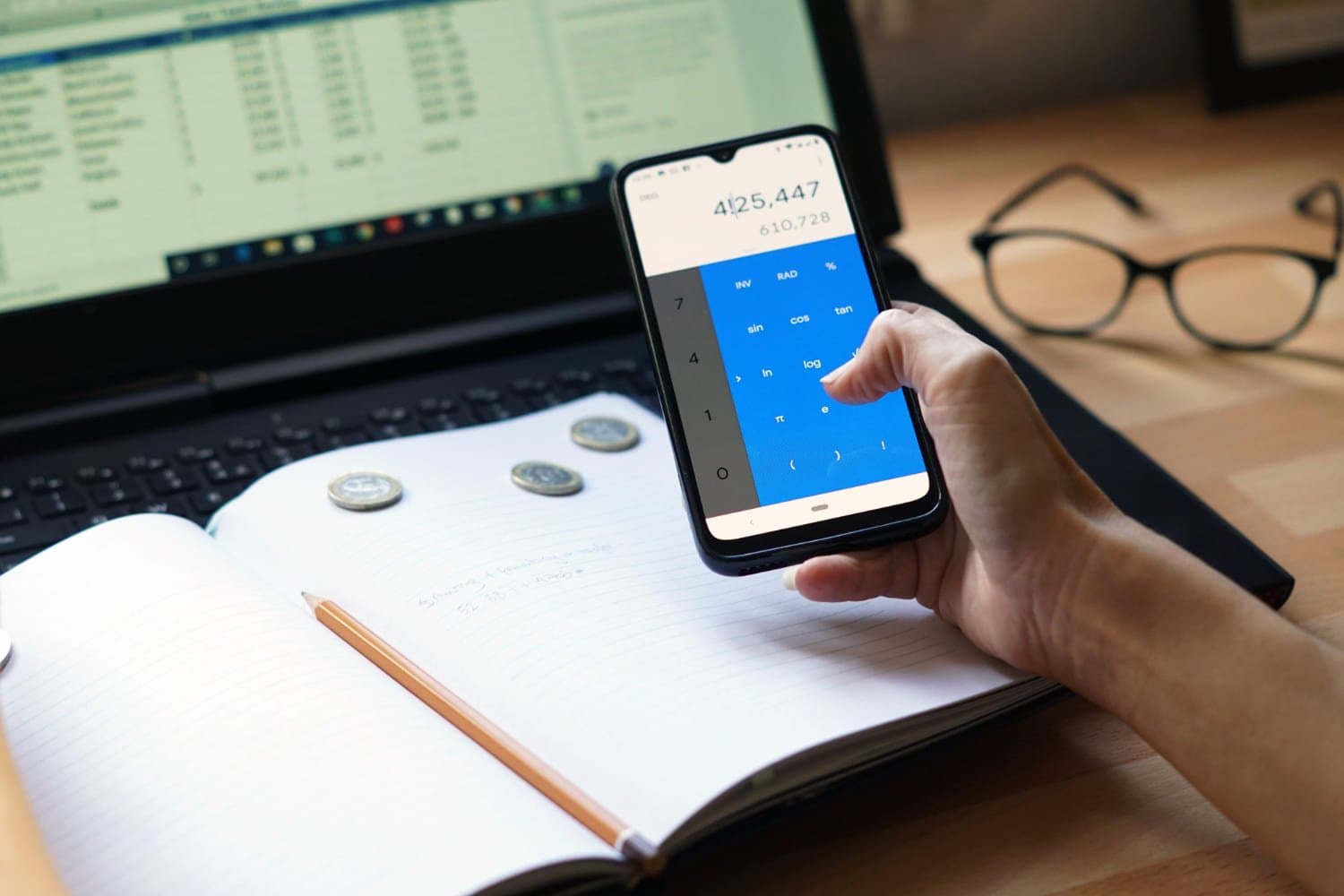 Saldo devedor do empréstimo consignado: tela de um notebook com planilha de fundo, enquanto à frente há um caderno em branco com um lápis e três moedas e uma mão segurando um celular enquanto utiliza a calculadora do dispostivo.