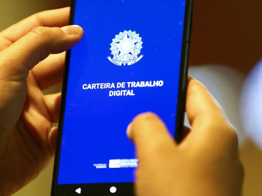 Uma pessoa branca mexe no celular e, na tela, é mostrado o aplicativo Carteira de Trabalho Digital.