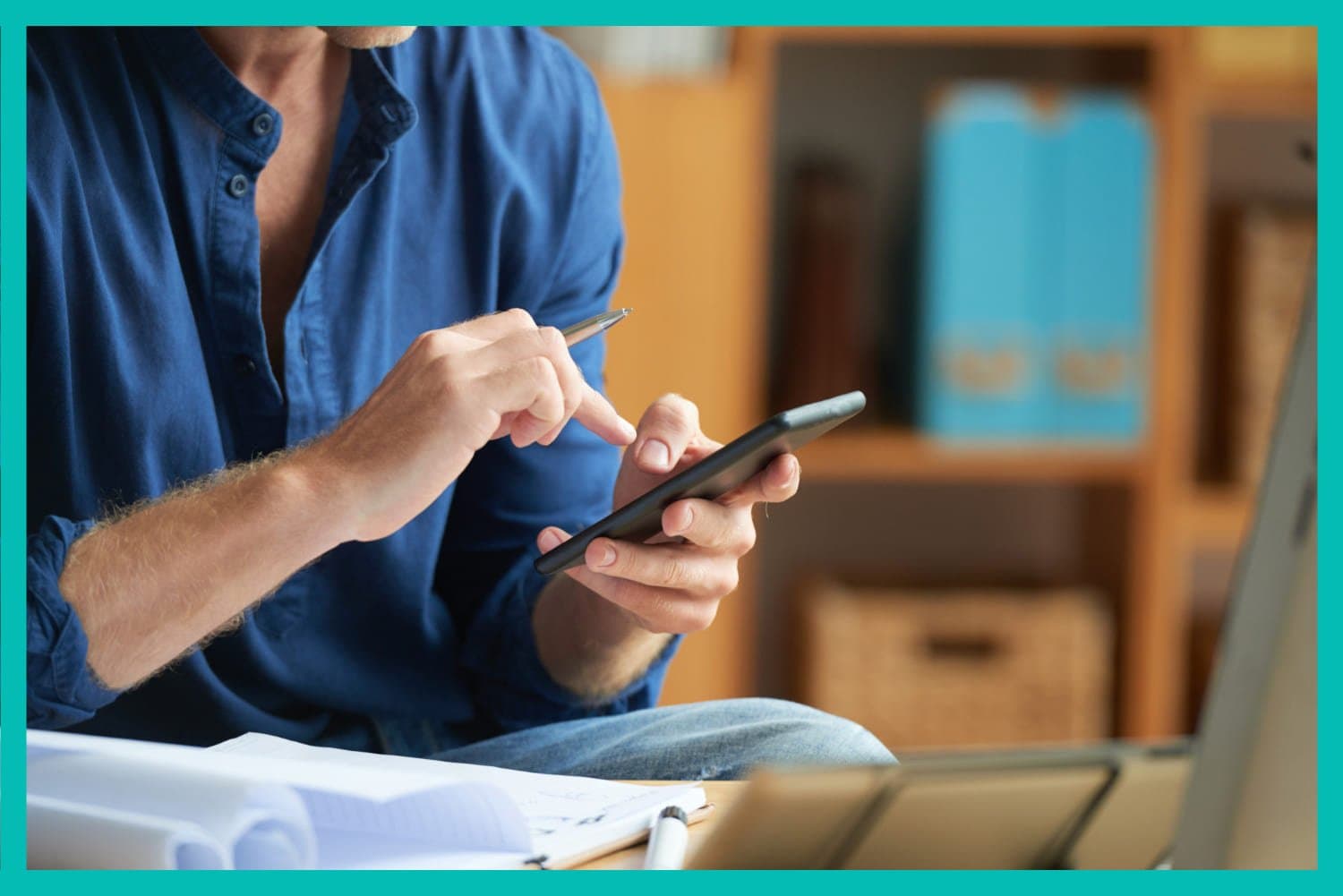Pix Parcelado: um homem de camisa azul mexe no celular. No fundo há algumas pastas azuis e uma caixa de madeira.