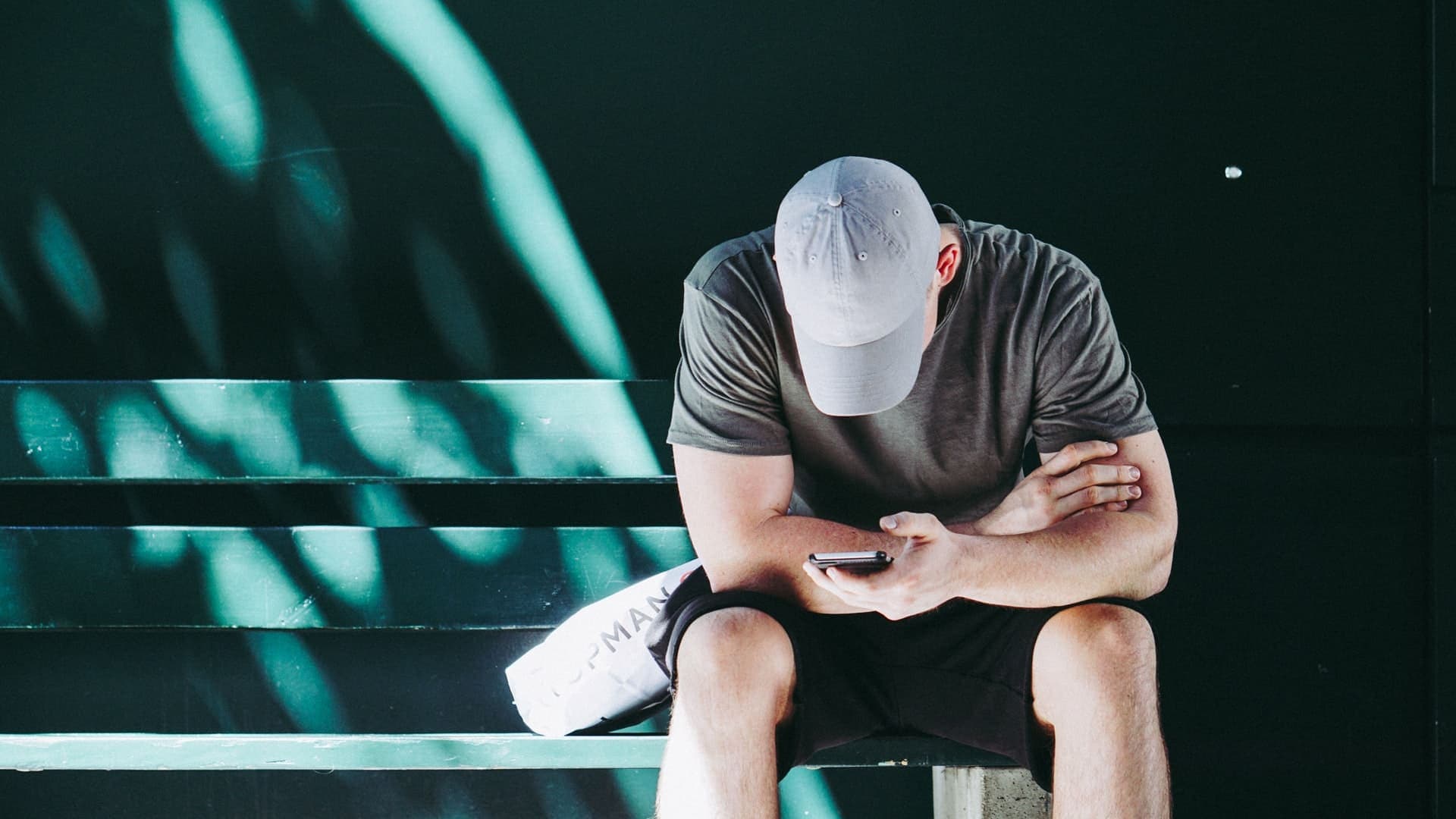Pix: um homem branco está sentado em um banco mexendo no celular. Ele veste camiseta, bermuda e boné.