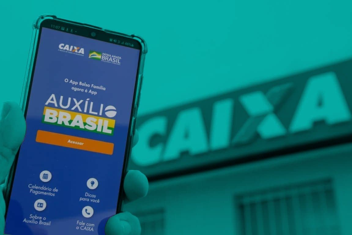 Auxílio Brasil: celular mostrando a tela de início do app Auxílio Brasil em frente a uma agência da Caixa
