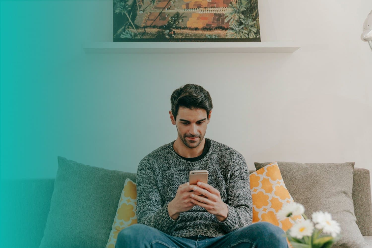 Cadastro Único: um homem branco está sentado num sofá cinza escuro , mexendo no celular. Atrás há algumas almofadas laranjas.