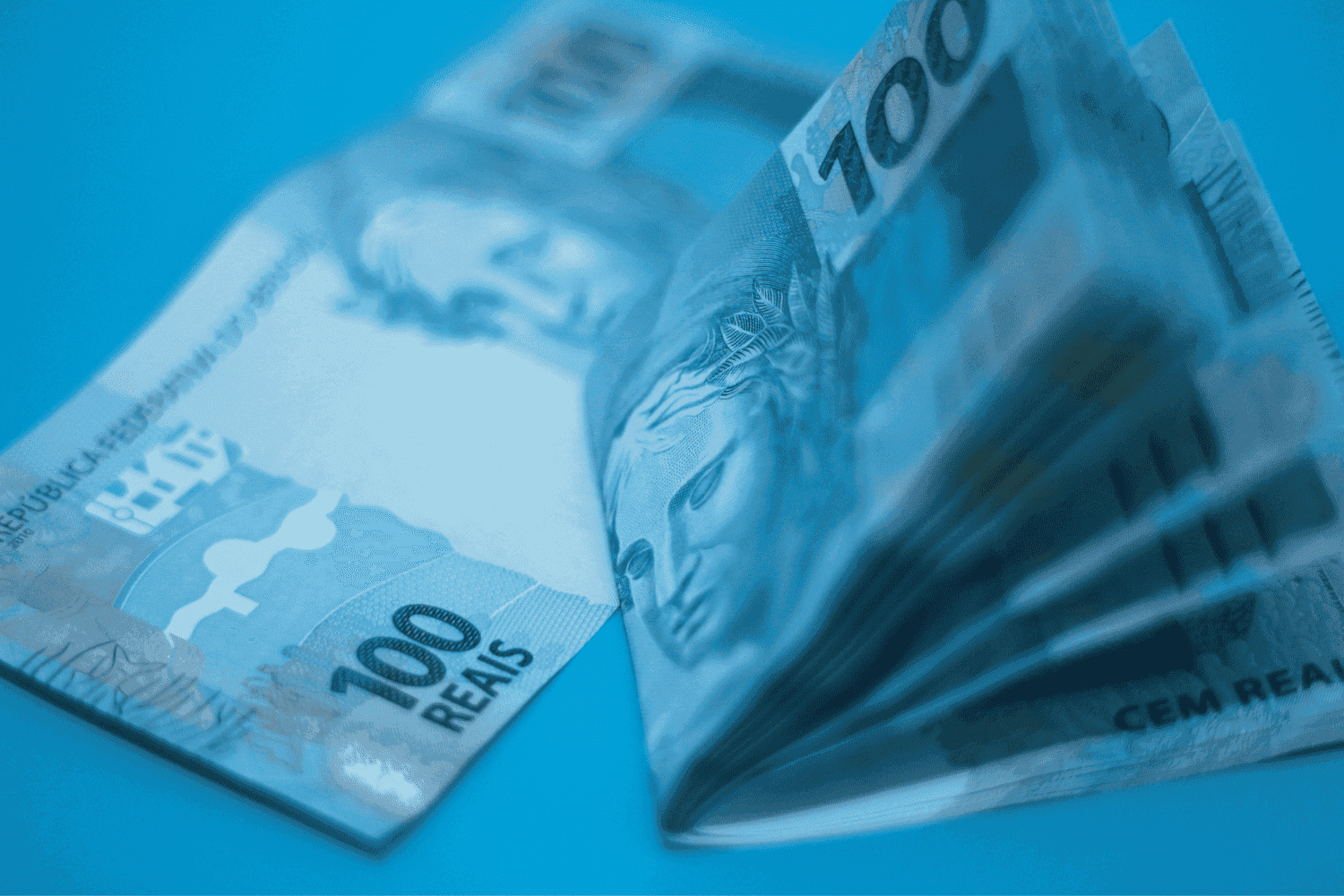 Auxílio Brasil: uma nota de cem reais está esticada numa superfície, enquanto outras notas estão dobradas.