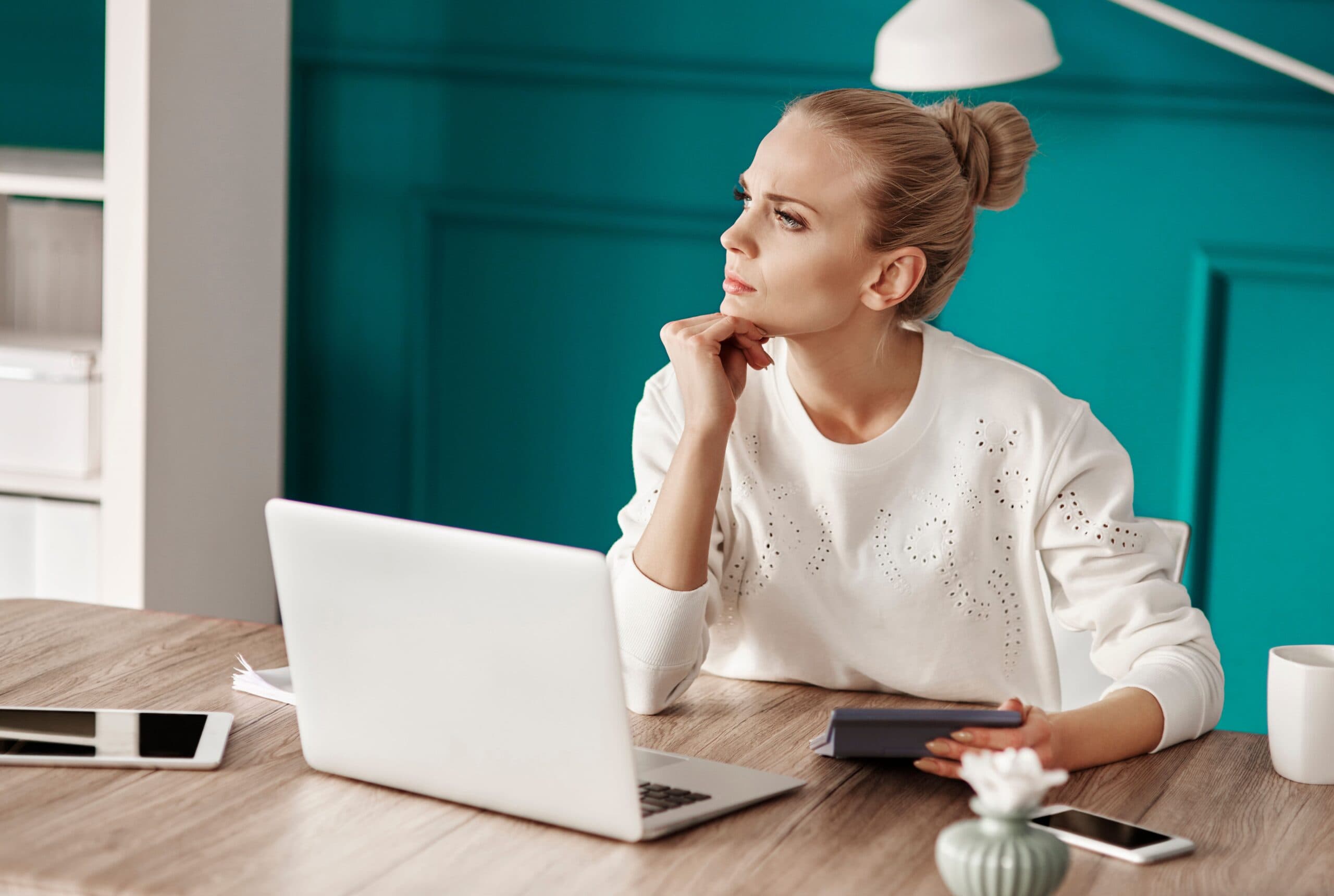 Empréstimo: uma mulher está sentada à mesa, refletindo. Em sua frente há um notebook e celular.