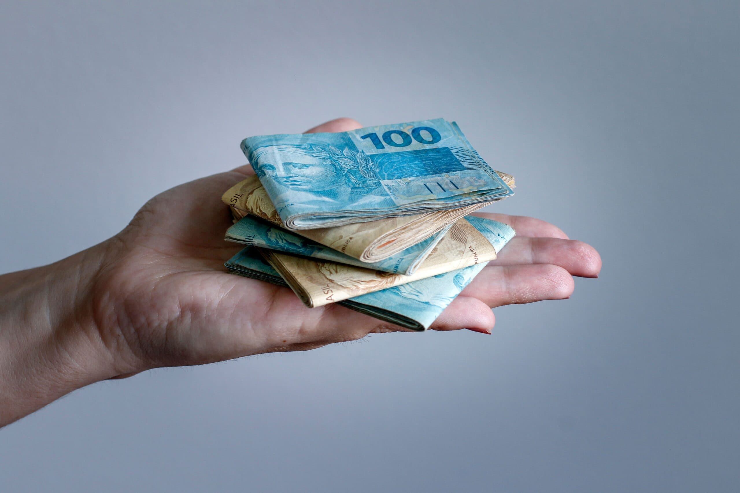 Saque-Aniversário: uma mão estendida equilibra diversas notas de cinquenta e cem reais.