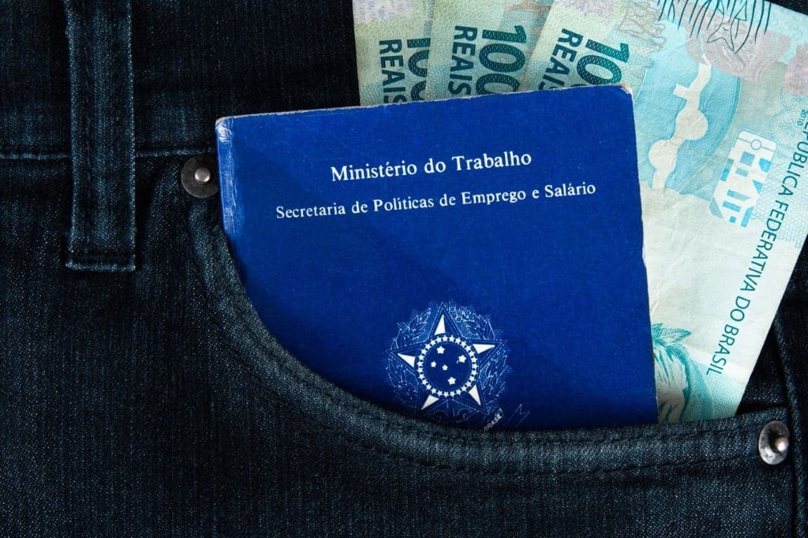 Seguro-Desemprego: três notas de cem reais e uma carteira de trabalho estão no bolso de uma calça jeans.