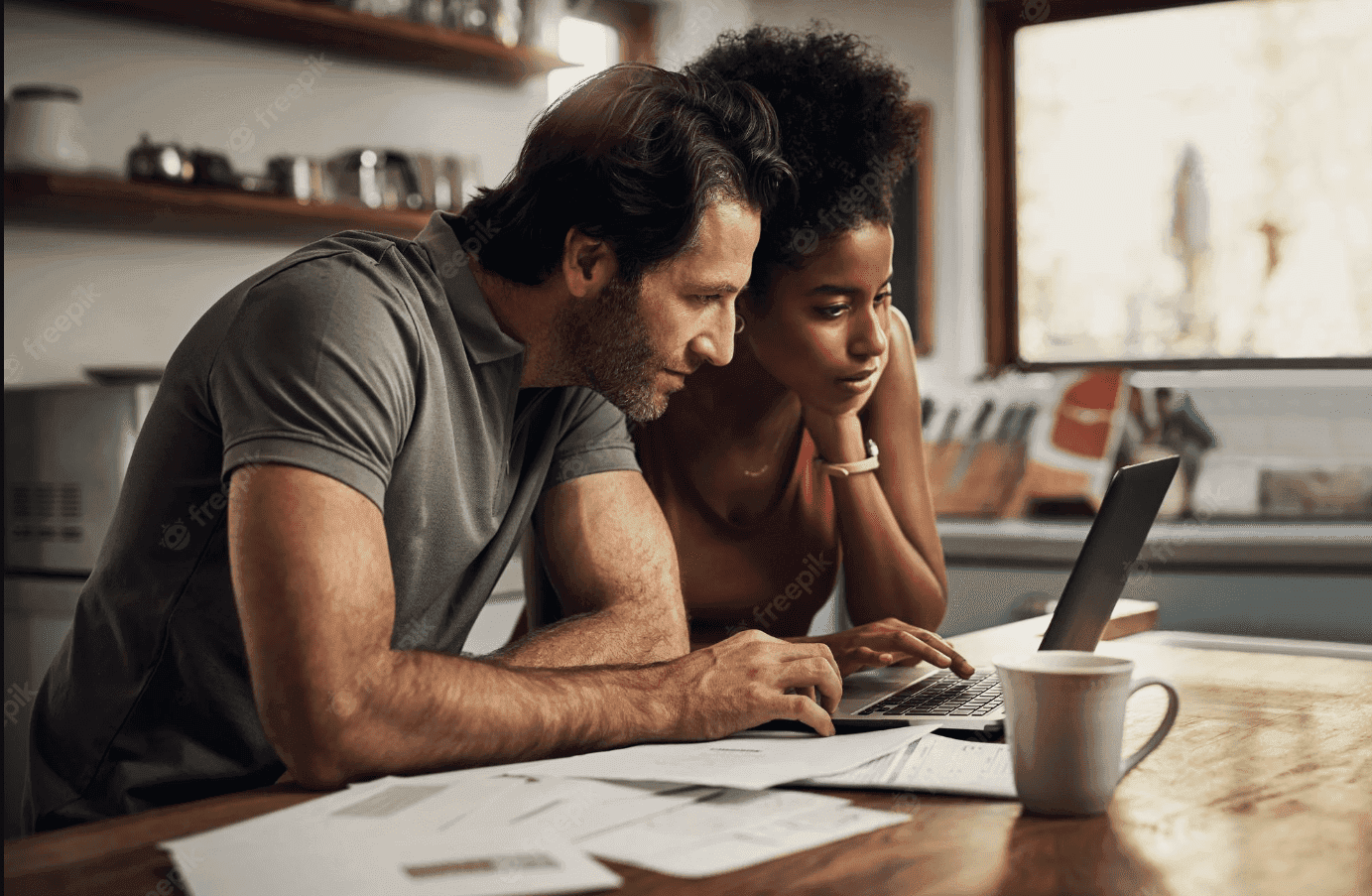 Conta conjunta: um casal está sentado à mesa, mexendo no notebook. Ao lado há uma xícara e alguns papéis.