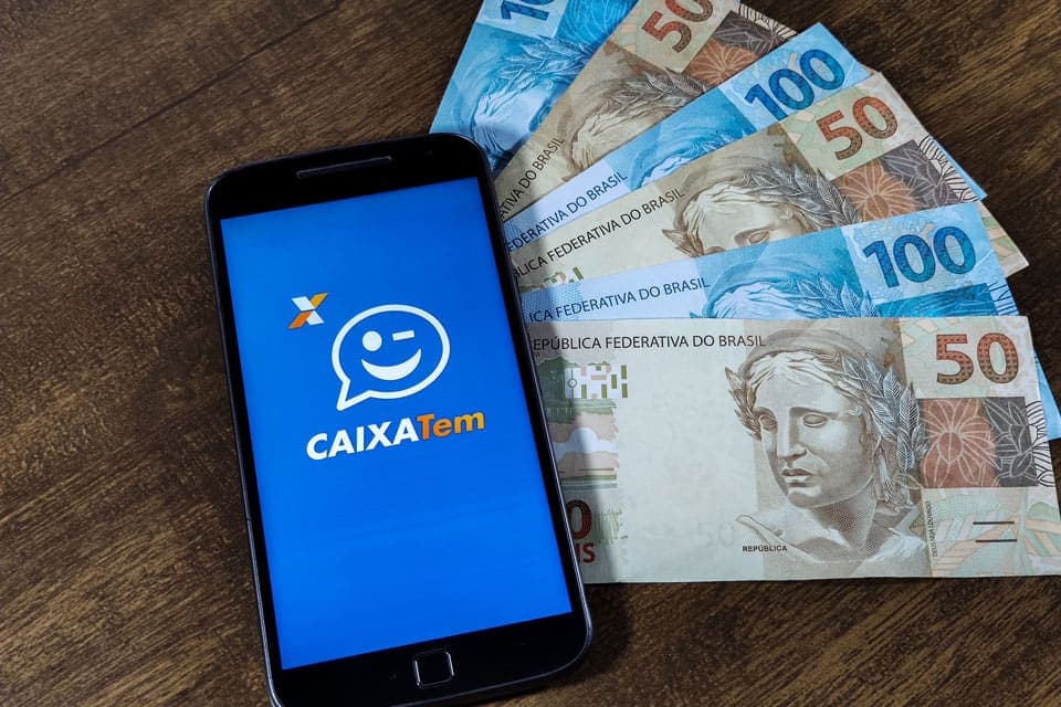 Empréstimo Caixa Tem para negativado: um celular possui o logo do Caixa Tem e, ao lado, há três notas de cinquenta reais e três notas de cem reais.