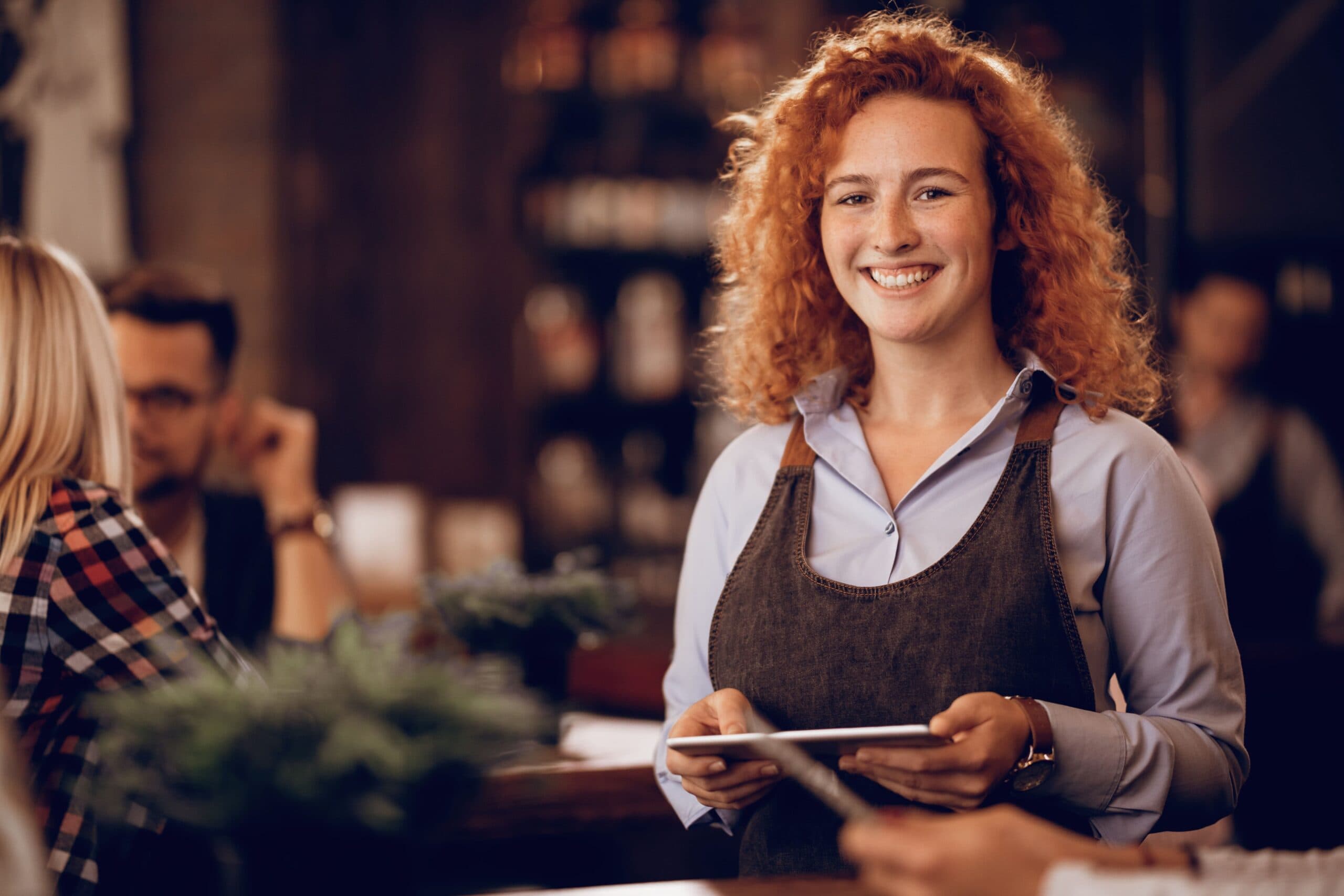 Empréstimo para mulher empreendedora: uma mulher ruiva, de cabelo cacheado, está em pé, segurando um tablet e sorrindo. Ela veste um avental.