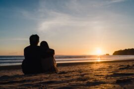 casal sentado na praia, olhando o pôr do sol (consórcio para lua de mel)