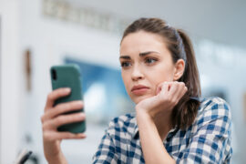 Golpes em aplicativo de pagamento: uma mulher está sentada, apoiando o rosto em uma das mãos e segurando um celular. Sua feição é de insatisfação.
