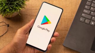 Pix no Google Play: uma pessoa segura um celular e, na tela, é mostrado o logo da Google Play. Atrás há uma planta e um notebook.