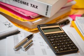 Imposto de Renda: imagem mostra papéis sobre uma mesa, uma calculadora ao lado de uma caneta e, no fundo, há um livro sobre impostos.