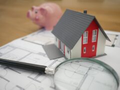 Financiar casa: miniatura de uma residência, uma caneta, um projeto arquitetônico, uma lupa e um cofre em formato de porquinho.