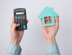 Crédito imobiliário: uma pessoa segura uma calculadora em uma mão e, na outra, a miniatura de uma residência em madeira.