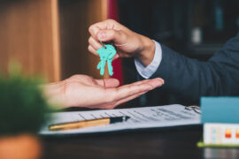 Refinanciamento de imóvel: uma pessoa entrega três chaves a outra pessoa. Na mesa há um contrato, uma caneta e decorações.