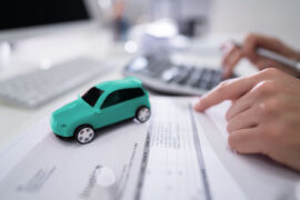refinanciar veículo: uma pessoa utiliza a calculadora e segura uma caneta, enquanto consulta dados em um documento. Ao lado há um veículo verde em miniatura e um computador.