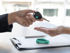Refinanciamento de veículo alienado: uma pessoa entrega a chave de um carro para outra. Abaixo, há um contrato, uma caneta e a miniatura de um veículo.