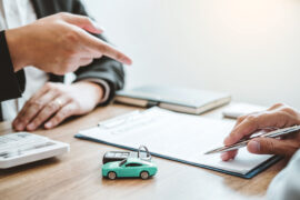 Financiamento ou consórcio: duas pessoas conversam a respeito de um contrato, que está na mesa. Ao lado, há uma calculadora, uma chave de carro e um veículo em miniatura.
