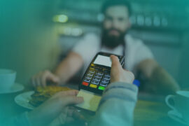 Uma pessoa está inserindo o cartão de crédito na maquininha de pagamento, enquanto o funcionário, de camiseta branca e avental escuro, aguarda.