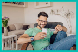 limite do cartão de crédito: homem sentado confortavelmente segurando o celular em uma mão e na outra um cartão.