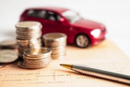 Financiamento: um veículo de brinquedo vermelho está atrás de um empilhado de moedas. à frente há uma caneta e abaixo um papel