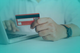 Um homem branco segura um cartão de crédito vermelho, enquanto digita no notebook. Ele veste uma camisa branca e tem um relógio no pulso.