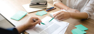 Financiamento: duas pessoas analisam um contrato. Ao lado há um bolo de notas, canetas e uma agenda.