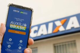 Auxílio Brasil: celular mostrando a tela de início do app Auxílio Brasil em frente a uma agência da Caixa