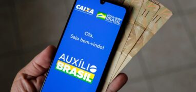 Auxílio Brasil: celular mostrando a tela de início do aplicativo Auxílio Brasil e notas de cinquenta reais atrás do celular