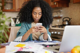 Empréstimo na conta de luz: uma mulher está sentada, mexendo no celular e, a sua frente, há um notebook e alguns papéis.