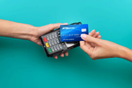 Uma pessoa aproxima o cartão de crédito da maquininha para realizar um pagamento. O fundo da imagem é verde.