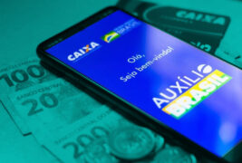 Auxílio Brasil: celular mostrando o logo do Auxílio Brasil na tela e apoiado sobre notas de vinte, cem e duzentos reais. Sobre a nota de duzentos há uma moeda de um real