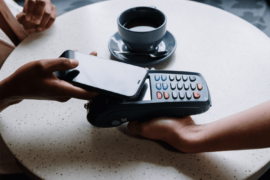 Cartão: uma pessoa realiza o pagamento através do celular, aproximando-o da maquininha. Na mesa, há uma xícara de café.