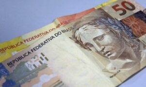 Auxílio Brasil: uma nota de vinte reais está abaixo de uma nota de cinquenta reais, ambas numa superfície lisa e branca.