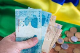Auxílio Brasil: notas de cinquenta e cem reais e moedas. Bandeira do Brasil ao fundo