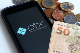 Pix Saque: um celular mostra o logo do Pix e, ao lado, há uma nota de cinquenta reais e moedas de cinco e vinte e cinco centavos e de um real.
