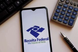 Empréstimo com garantia: a tela de um celular mostra o logo do aplicativo Meu Imposto de Renda, da Receita Federal. Ao lado há uma caneta, uma calculadora e um teclado.